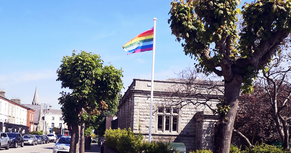 A building surrounded by trees on a quiet street with a rainbow flag on a pole outside