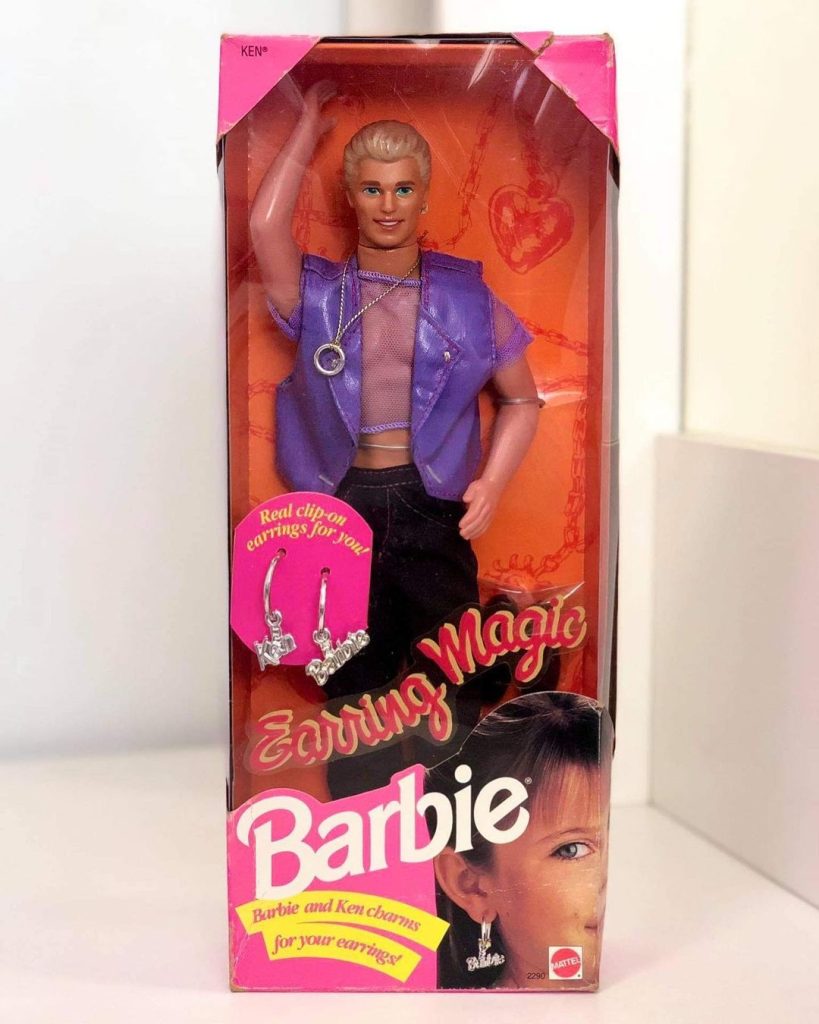 An Earring Magic Ken doll, often known as Gay Ken, dressed in a purple mesh shirt, purple faux-leather jacket and black pants, in its cardboard 'Barbie' packaging.