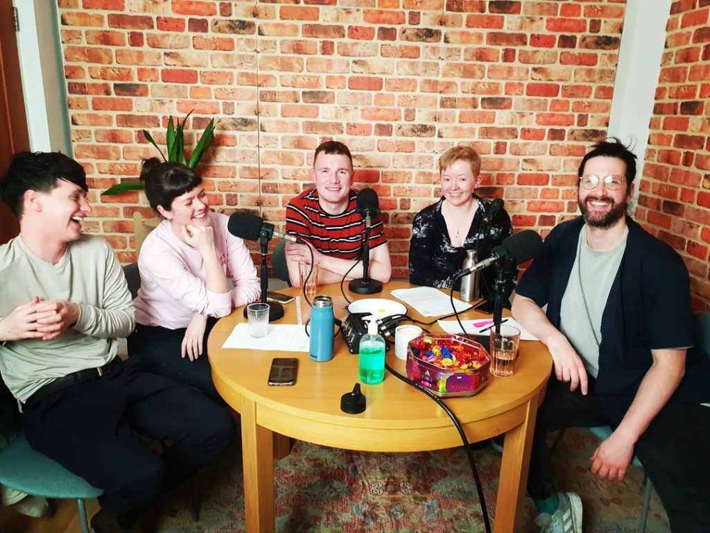 Five people seated around a table with microphones, recording the new podcast from ShoutOut.