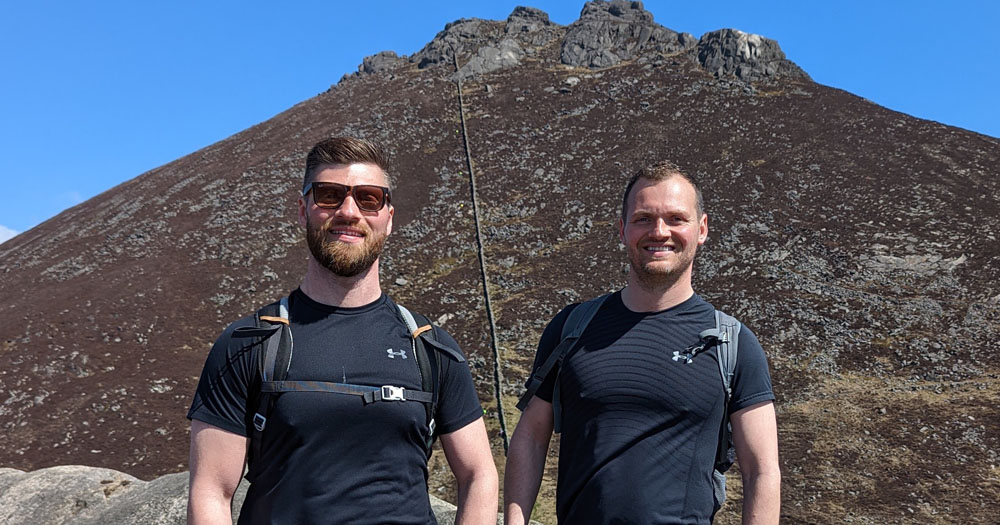 The photograph shows Luke and Dean, two of the members of Out & About Hikers. The two men are wearing black tshirts and backpacks. They are standing in front of the peak of the Slieve Binnian mountain.