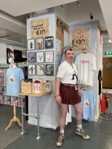 Colm Molloy at Dublin Pride Hub in front of their artwork including prints, t-shirts, and badges.