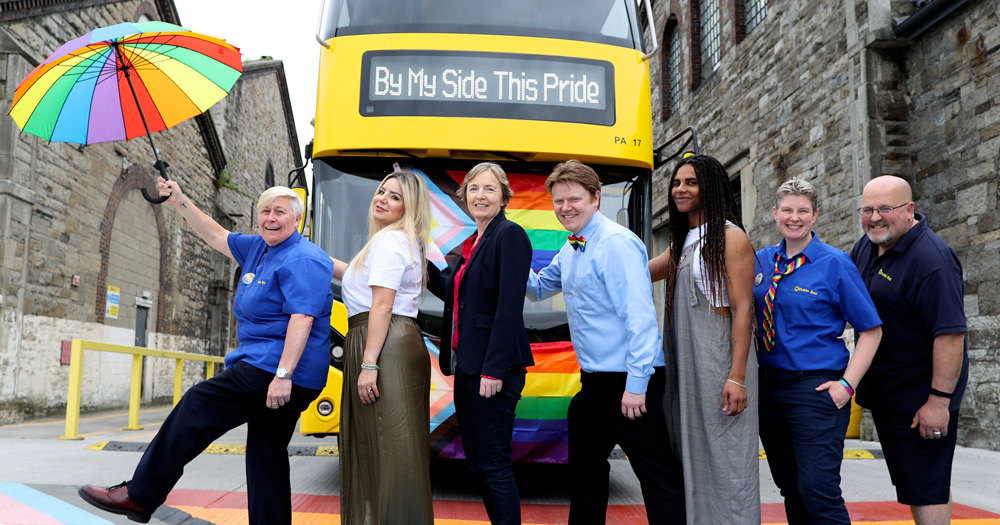 Image of the ambassadors for the Dublin Bus Pride parade. They are posing in front of a Dublin Bus on a rainbow flag crosswalk.