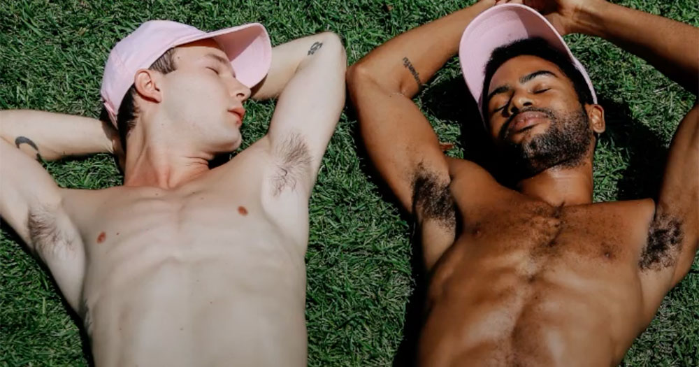 Two people laying on the grass with pink baseball caps on. Image from a Grindr advertisement for the new filter for sides on Grindr.