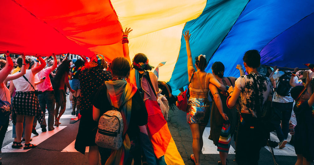 WHO have issued statements in support of LGBTQ+ Pride in light of monkeypox outbreak. The photograph shows young people walking under a giant Pride flag from behind.