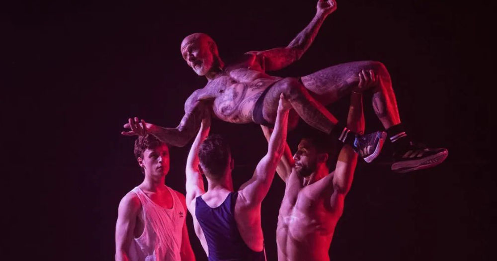 The photograph shows a scene from the dance performance Party Scene. In the image 2 men are lifting another man who is only wearing shorts and trainers. A fourth man stands looking at them.