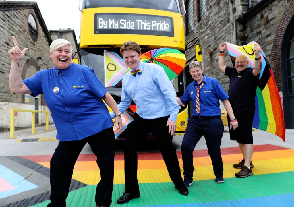 Individuals part of the Dublin Bus Pride campaign walking along a Pride flag crosswalk. 