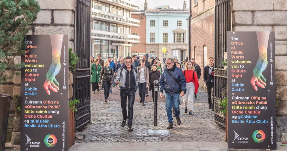 The photograph shows Tonie Walsh bringing a group on his Lavender Walk for Culture night. The group are coming through the arch of Dublin Castle with Culture Night poster on either side