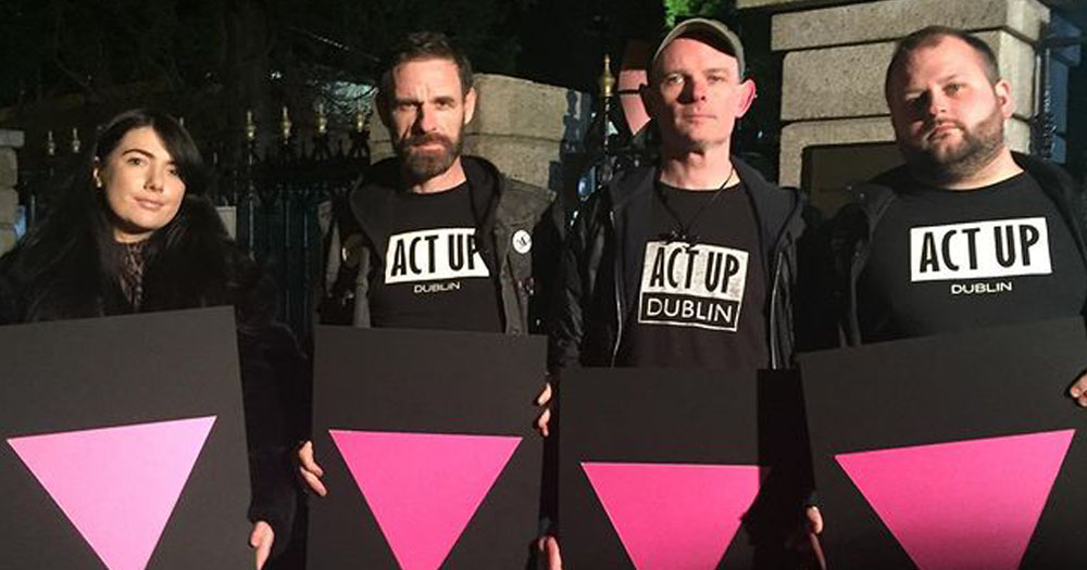 The photograph shows four members of ACT UP Dublin who are calling for Monkey Pox vaccinations. In the photograph the group are demonstrating at a night protest all wearing black with white wording reading ACT UP Dublin. They are all holding placards with a pink triangle against a black background.