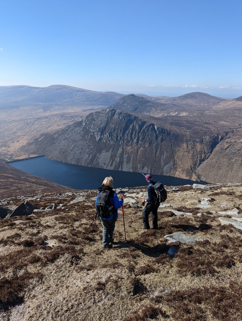 The photograph shows two hikers looking and pointing towards a lake. They are standing on a hill above the lake facing other mountains on the other side of the lake.
