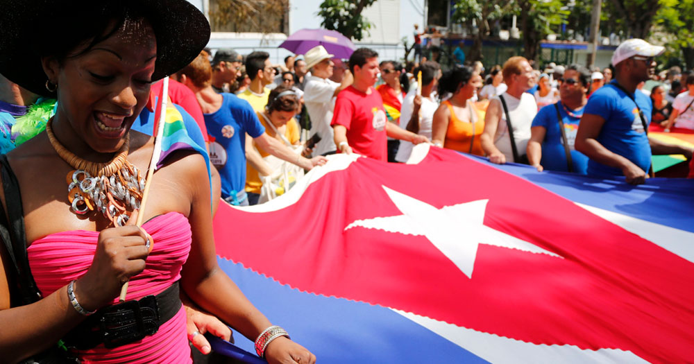 Cuba has announced that it is to hold a referendum on same-sex marriage. The photograph shows a person in a pink dress waving a miniature Pride flag over their shoulder with the other hand holding a giant Cuban flag. In the background a crowd of other people are also holding the Cuban flag.