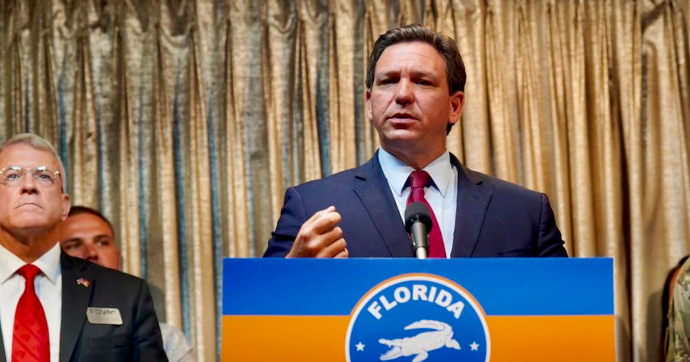 Photograph of Governor Ron DeSantis who passed the 'Don't Say Gay' law last March. DeSantis is standing in front of a podium with the Florida emblem on it. He is wearing a blue red tie.