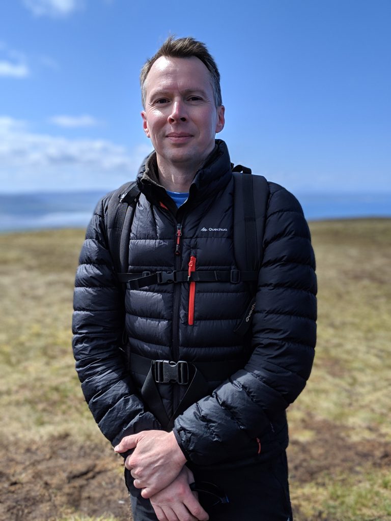 The photograph shows Neal Lester, the founder of Out & About Hikers. Neal is looking straight into the camera wearing a padded hiking jacket. The photograph is taken in a mountain setting.