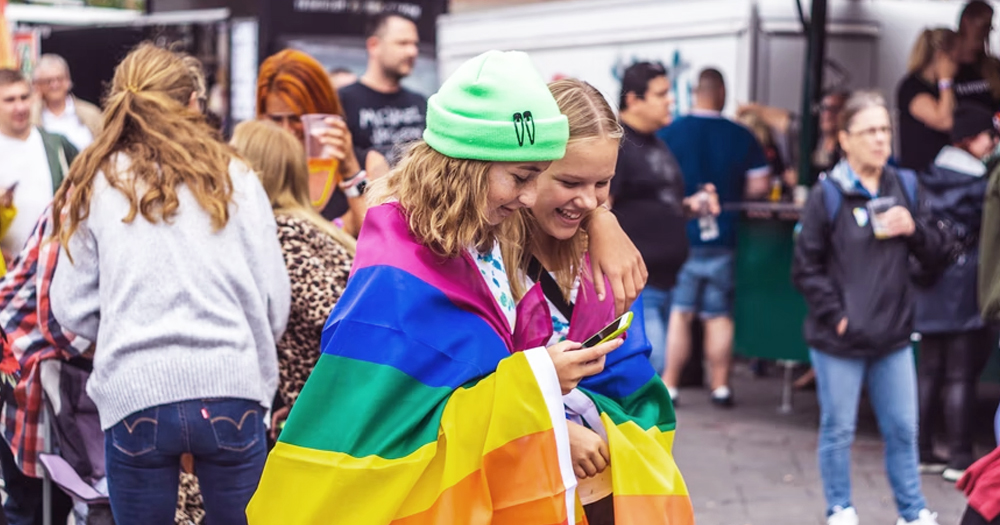 This article is about Pride events happening this week in Ireland. In the picture, two girls hugging each other and wearing a rainbow flag, with other people in other background.