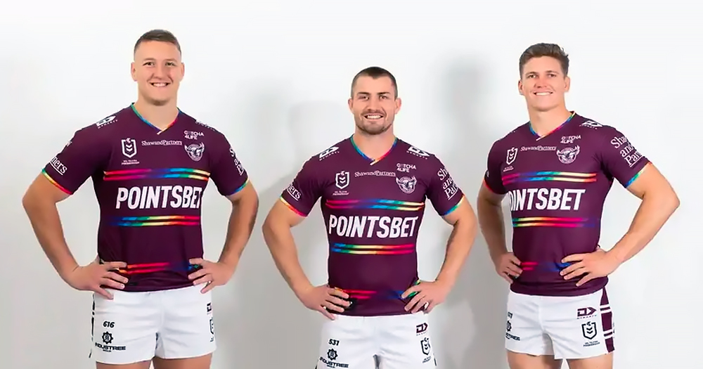 The Photograph shows three players from the Australia rugby league team Manly Sea Eagles wearing the new 'Everyone in League' jersey. the jersey is maroon colour with two bands of Pride colours across the centre above and below the sponsors name of Pointsbet.