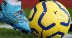 Homophobia is still rife in male sport. This photograph shows a close up of a blue soccer boot about to kick a yellow football. The boot is laced with rainbow colour laces.