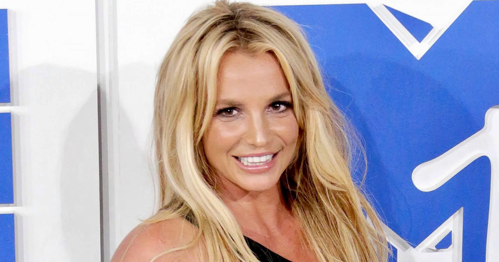 An image of pop singing icon Britney Spears posing on a red carpet. She is smiling against a white and blue background.