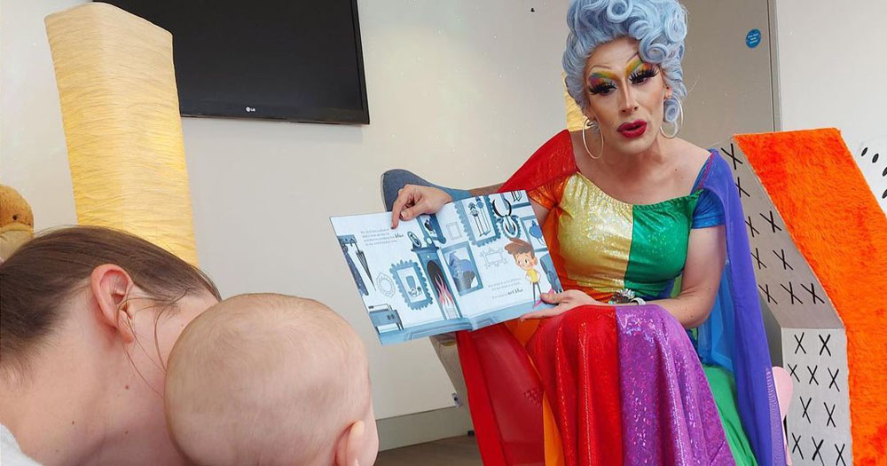 The photograph shows drag artist Cherrie Ontop reading a book to a baby. Cherrie is wearing a rainbow coloured dress with a light blue curly wig done in an up style. She also has rainbow coloured eyeshadow.
