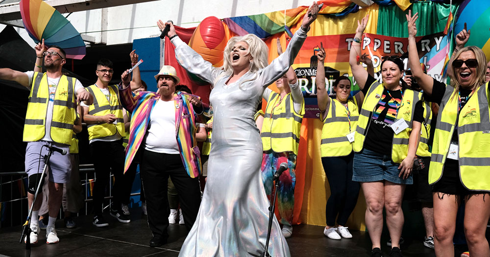Tina D. Parton celebrating at Clonmel Pride in front of a big Pride banner and a group of people wearing high-vis vests