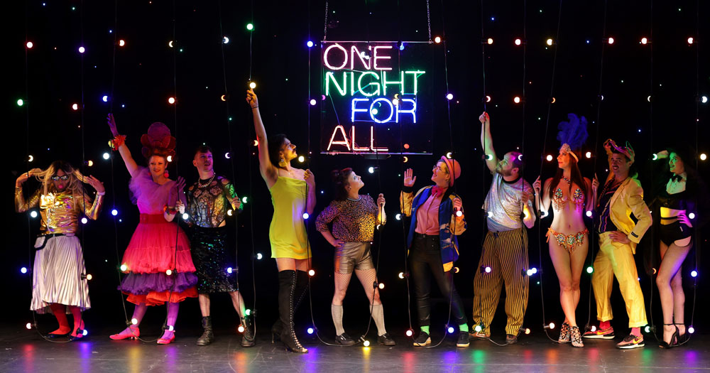 The photograph shows ten people lined up in sparkly party clothes. They are looking up at a neon sign which reads 