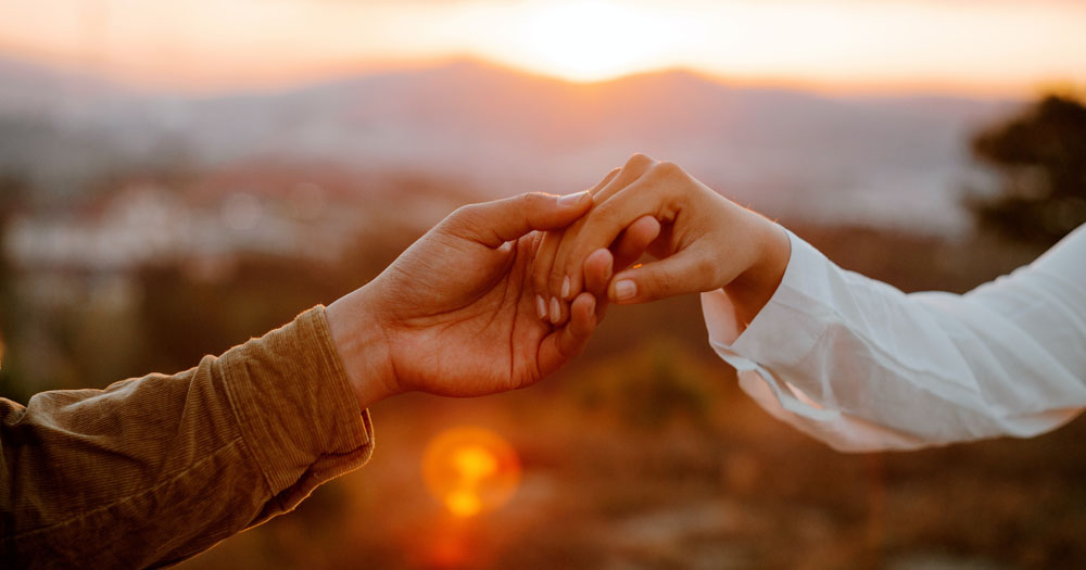 The photograph shows two hands holding in front of a sunset. The article explores age in relation to dating in the LGBTQ+ community.