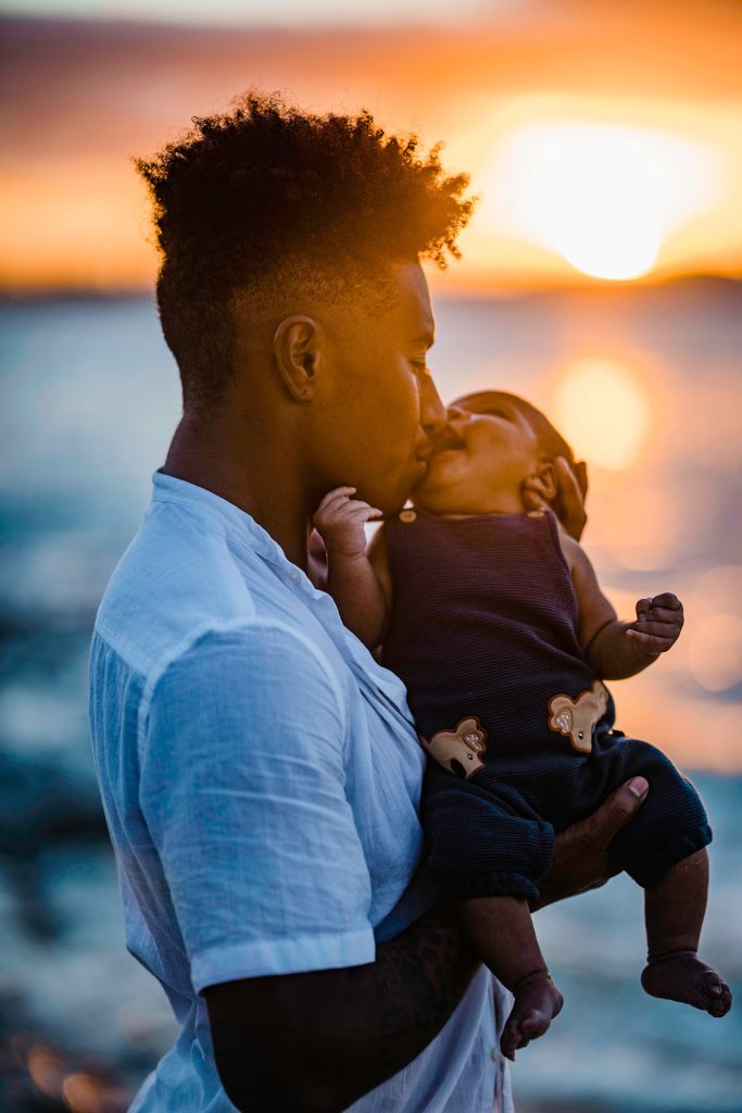 Photo of Ellia Green, who is the first Olympian to come out as a Trans man, holding his daughter with a sunset in the background.