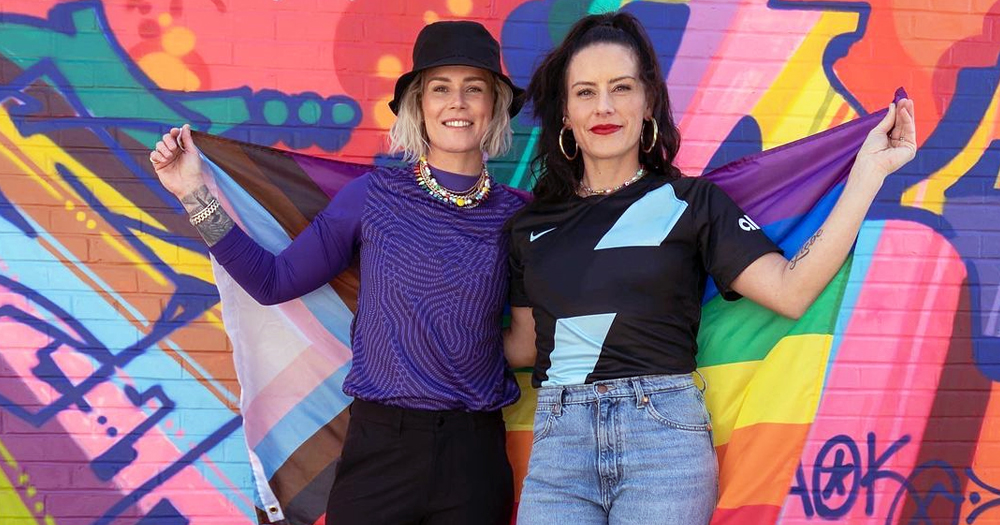 Photo of Ashlyn Harris and Ali Krieger, who just adopted a second child, holding a progress Pride flag with a colorful background.