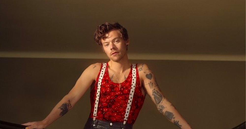 A photo of Harry Styles. He is leaning against a banister and wearing a red tank top with white suspenders.