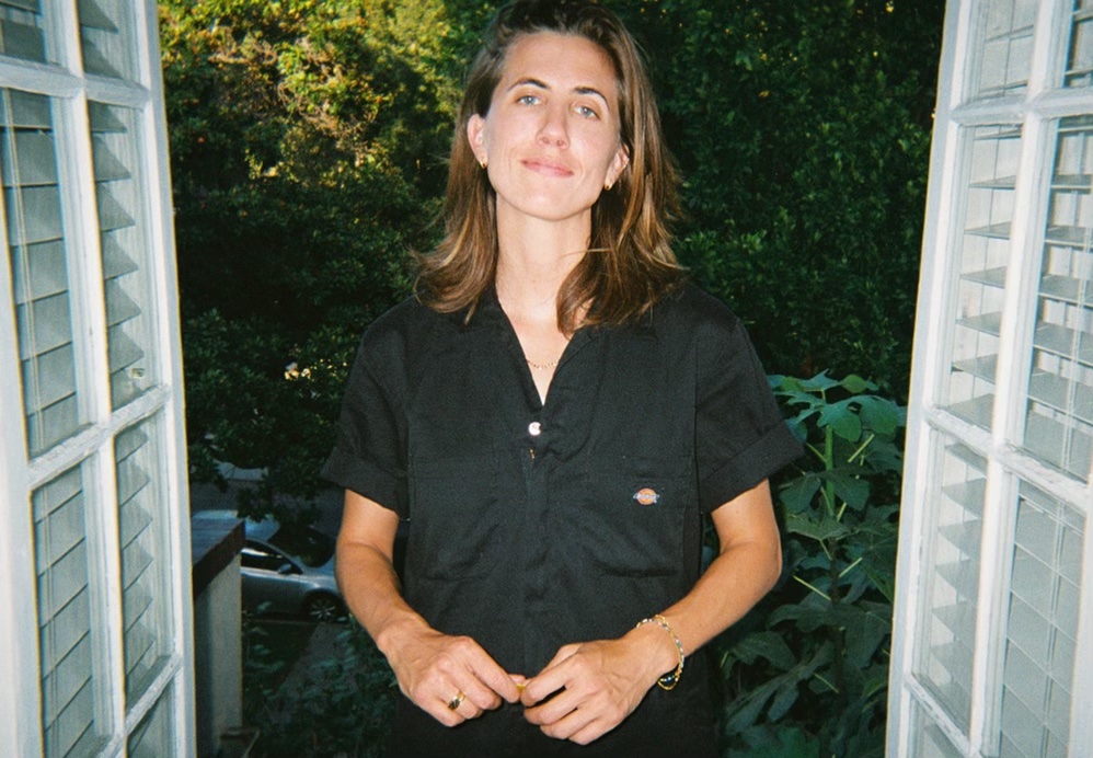 An image of E.R Fightmaster. They are wearing black overalls and standing in front of trees.