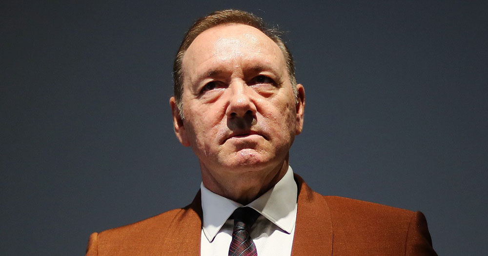 The image shows a head and shoulders portrait of Kevin Space, the House of Cards actor who has been forced to pay $31 million in damages. The photograph is taken front on looking up at Spacey who is wearing a brown blazer with a white shirt and black and red tie.