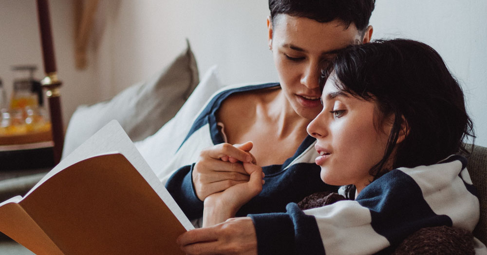 A photograph of two women cuddling while reading an LGBTQ+ Dictionary.
