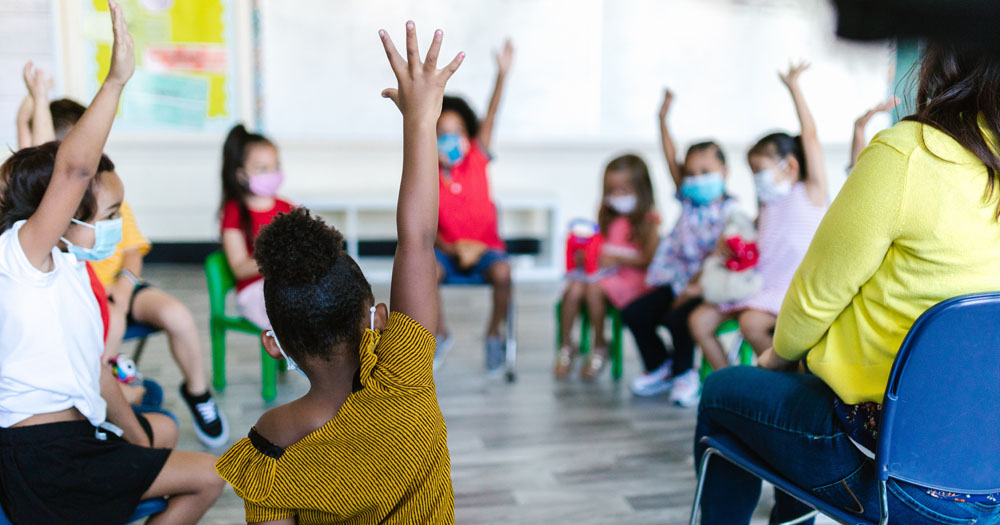 The photograph shows children in a classroom raising their hands. A child has been excluded from a school in Louisiana because she has been adopted by a same-sex couple.
