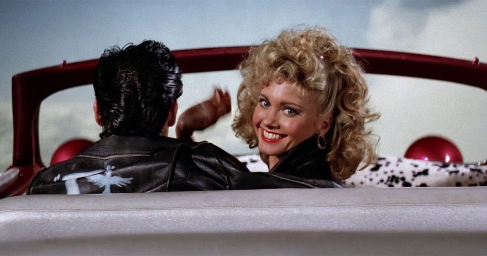 an image of Olivia Newton-John as Sandy in the iconic film Grease. She is sitting in a car with John Travolta and she is looking back and smiling at the camera.
