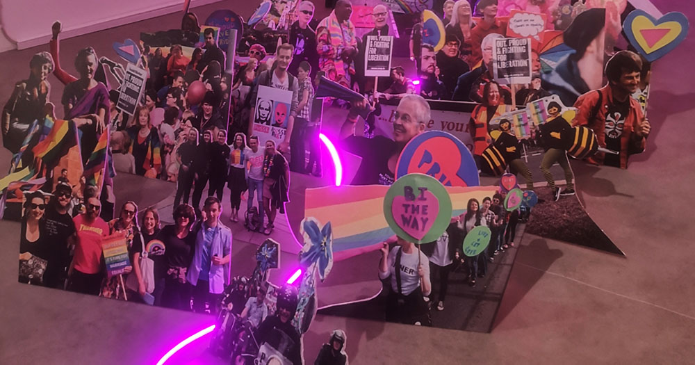 The photograph shows a series of images cut out from photographs of Foyle Pride parades for the RFR exhibition at Void Gallery in Derry. There is a fluorescent strip light running through the middle of the cutouts.