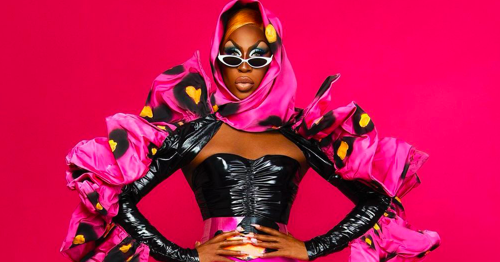 Shea Couleé poses in front of a pink background.