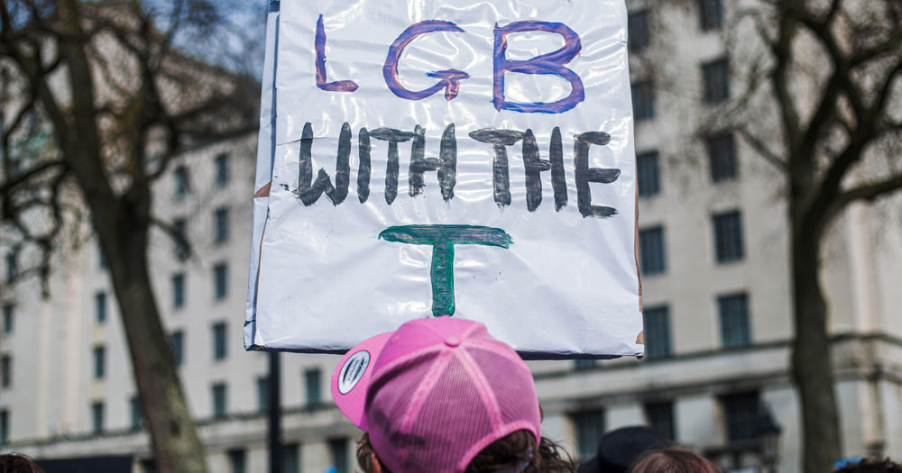 A person wearing a cap seen from behind holding a placard that reads 'No LGB without the T'