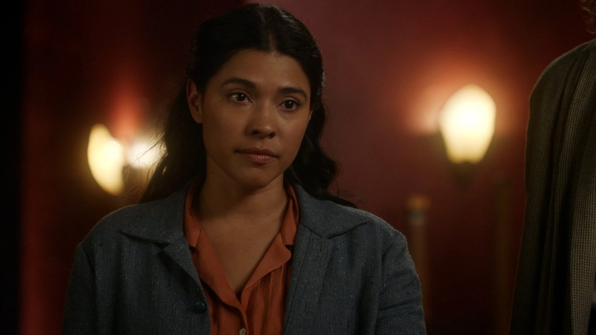 Asexual character Esperanza Cruz from Legends of Tomorrow stands against a warm brown background with a concerned look on her face.