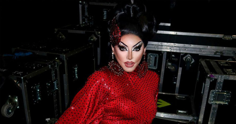 The image shows drag performer Cherry Valentine wearing a red dress with a high black wig.