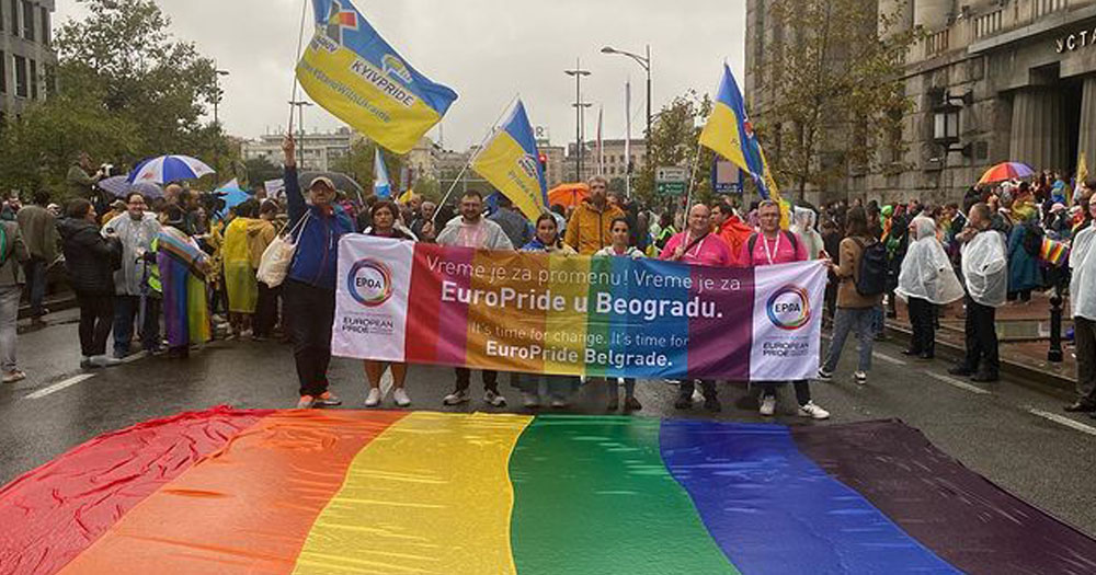 The image shows the start of the EuroPride 2022 parade in Belgrade. Marchers are standing behind a banner in front of a rainbow flag which is lying on the ground in the rain.