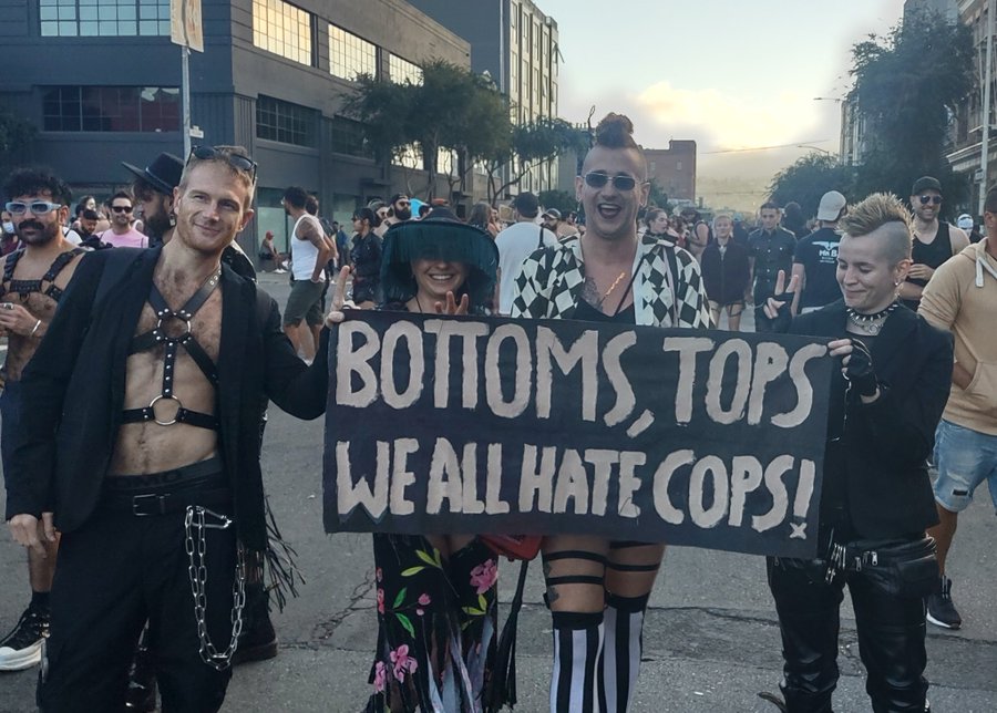 Four people holding a sign reading 'BOTTOMS, TOPS WE ALL HATE COPS!'