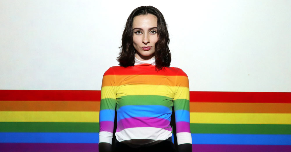 BeLong To have released a new survey investigating LGBTQ+ mental health in Ireland. The image shows a woman wearing a rainbow coloured sweatshirt standing in front of a wall with a rainbow.