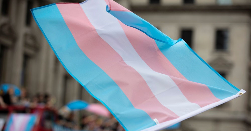 The image shows a trans flag waving in the wind. Montana has banned Trans people from changing their gender marker on their birth certificates.
