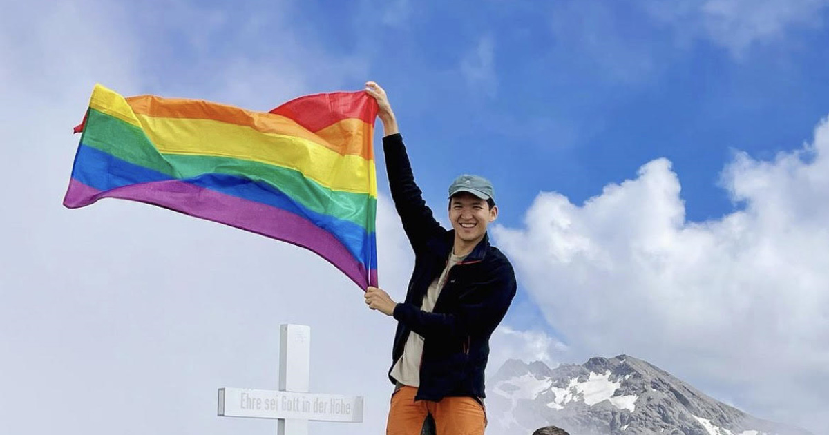 A man is holding a pride flag at the top of a mountain.