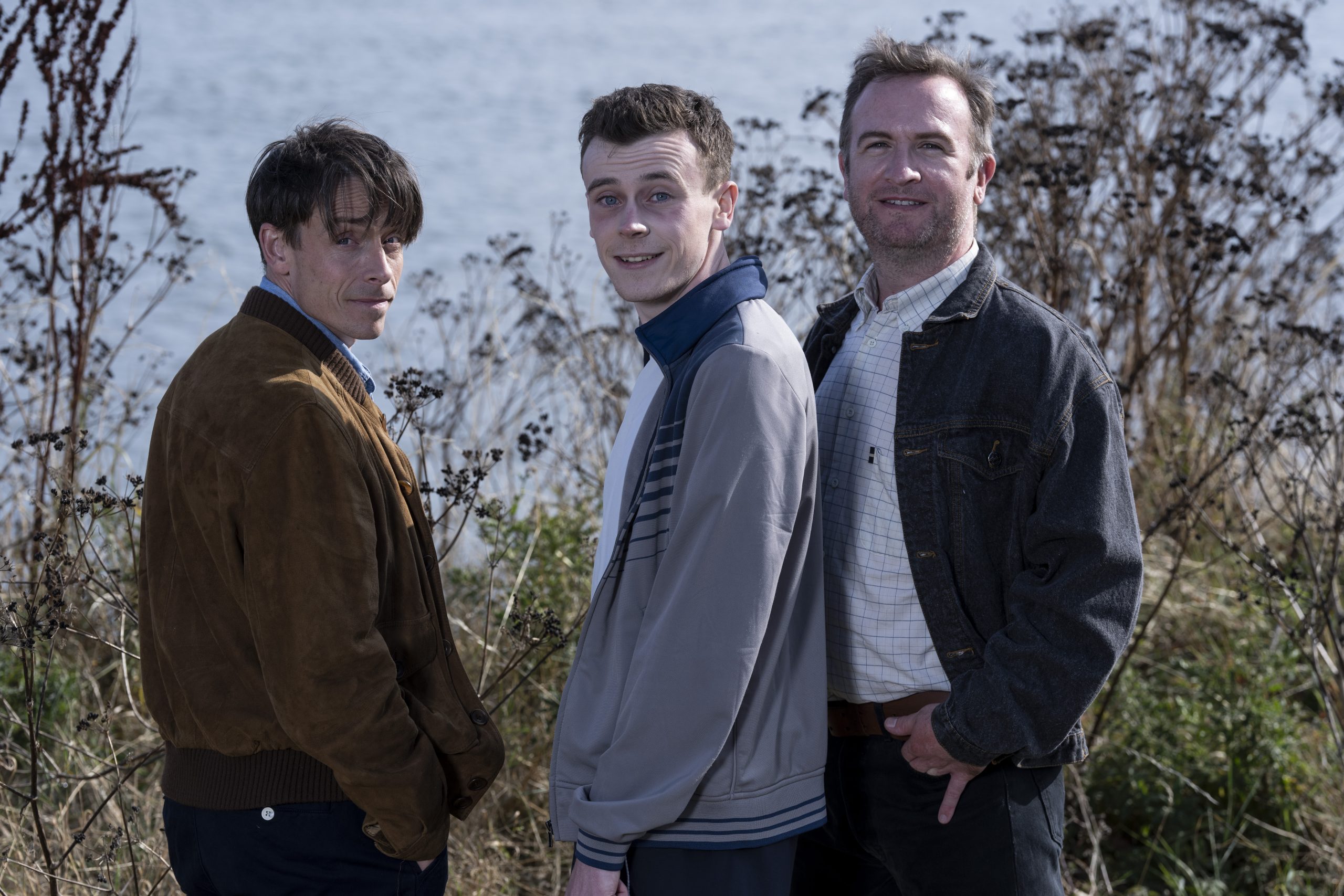 A photo of three of the actors in The Blackwater Lightship. They are outside amongst trees and turned around towards the camera