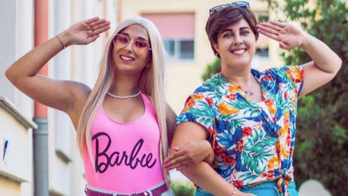 One girl wears a pink body suit with the words Barbie printed on the front. She links arms with another woman who is dressed as Ken, Barbie's boyfriend. She is wearing a tropical print shirt.