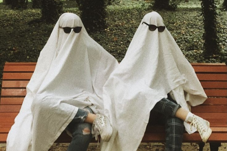 Two people sit on a bench with a bedsheet and sunglasses over them, a perfect last minute Halloween costume