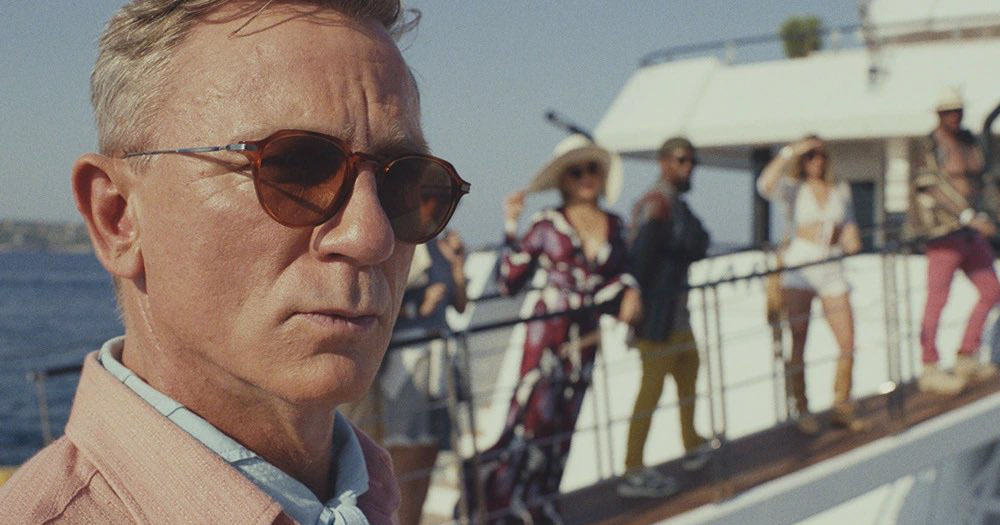 A still from the film 'Glass Onion: A Knives Out Mystery', showing a close up head shot of actor Daniel Craig wearing sunglasses with a gangway of people boarding a boat in the background.