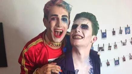 Two men dressed up in a Halloween couple's costume as comic book characters Harley Quinn and the Joker. They are both smiling and Harley Quinn is on Joker's back.