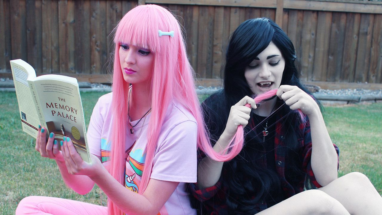 Two people are dressed as Marceline and Princess Bubblegum from the cartoon show Adventure Time. Bubblegum is reading a book while Marceline is trying to take a bit of Bubblegum's hair.