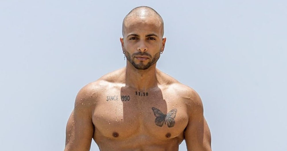 Photograph of Mr Gay World 2022. The image is from the chest up. He is topless with a butterfly on his left chest muscle and words tattooed around his neck. He has a shorn head with a thin beard.