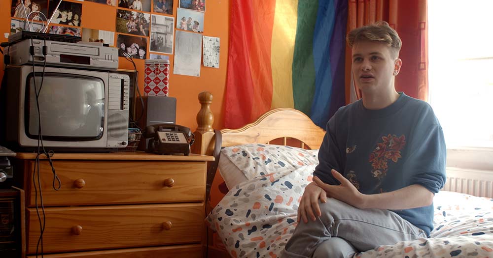 Luke Faulkner sits in front of pride flag. This image was taken from his Irish LGBTQ+ songwriter documentary